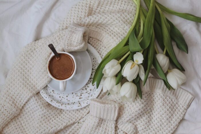 flowers and cup of cacao placed on knitted cardigan on bed