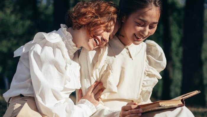smiling women in old fashioned clothing reading book on picnic blanket